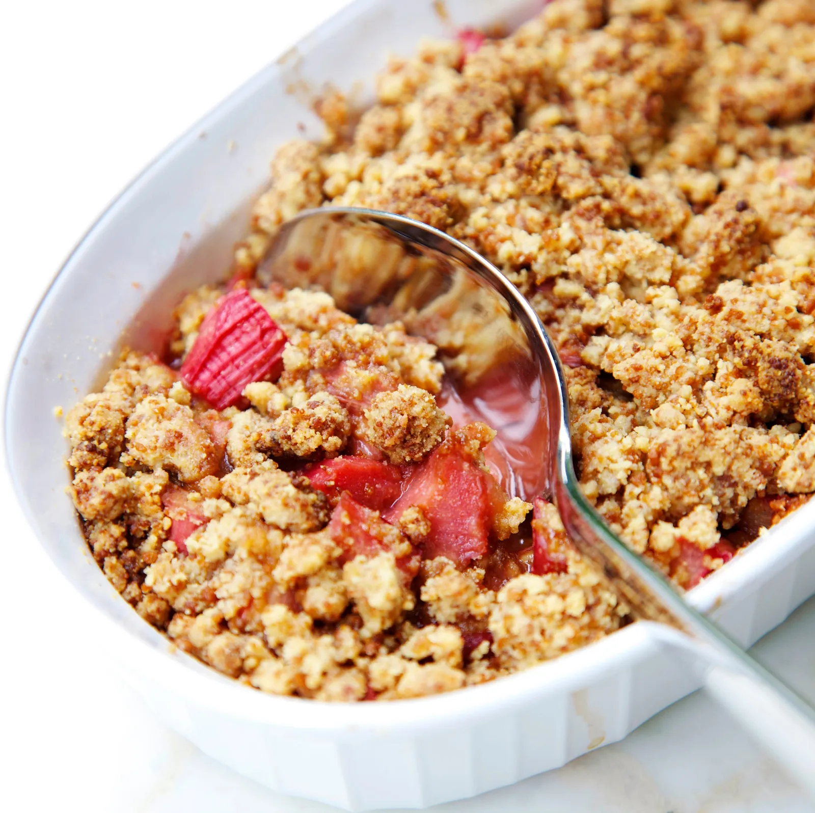 Crumble in weißer Form, ein Löffel bricht die Streusel auf und legt rosa Rhabarber frei